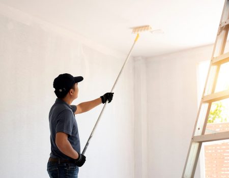 Asian rear view of a male painter drawing a wall with paint roller and a separate tank from a large empty space with wooden stairs.